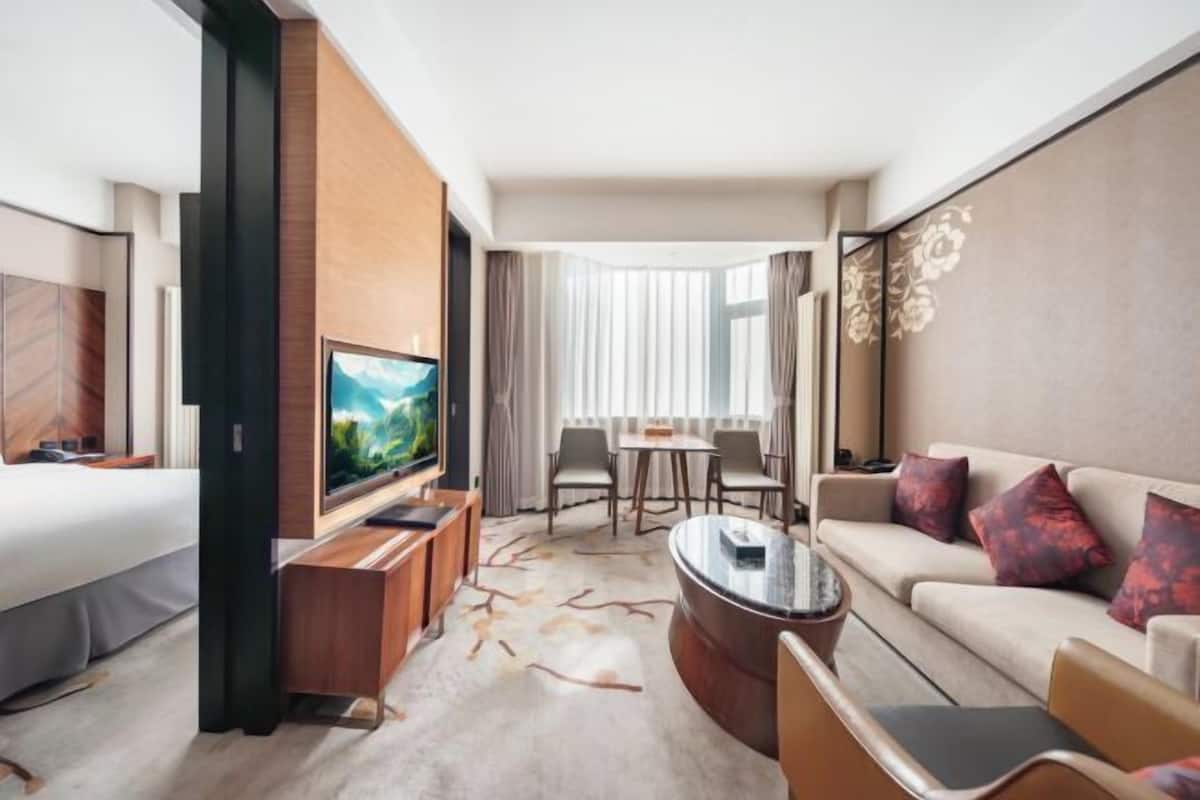 Una habitación de hotel moderna con una cama grande, un televisor, una zona de estar con una mesita de café y una mesa de comedor con sillas.