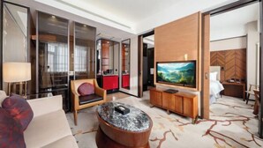 A modern hotel room with a sofa, a wooden coffee table, a TV, and a large window.