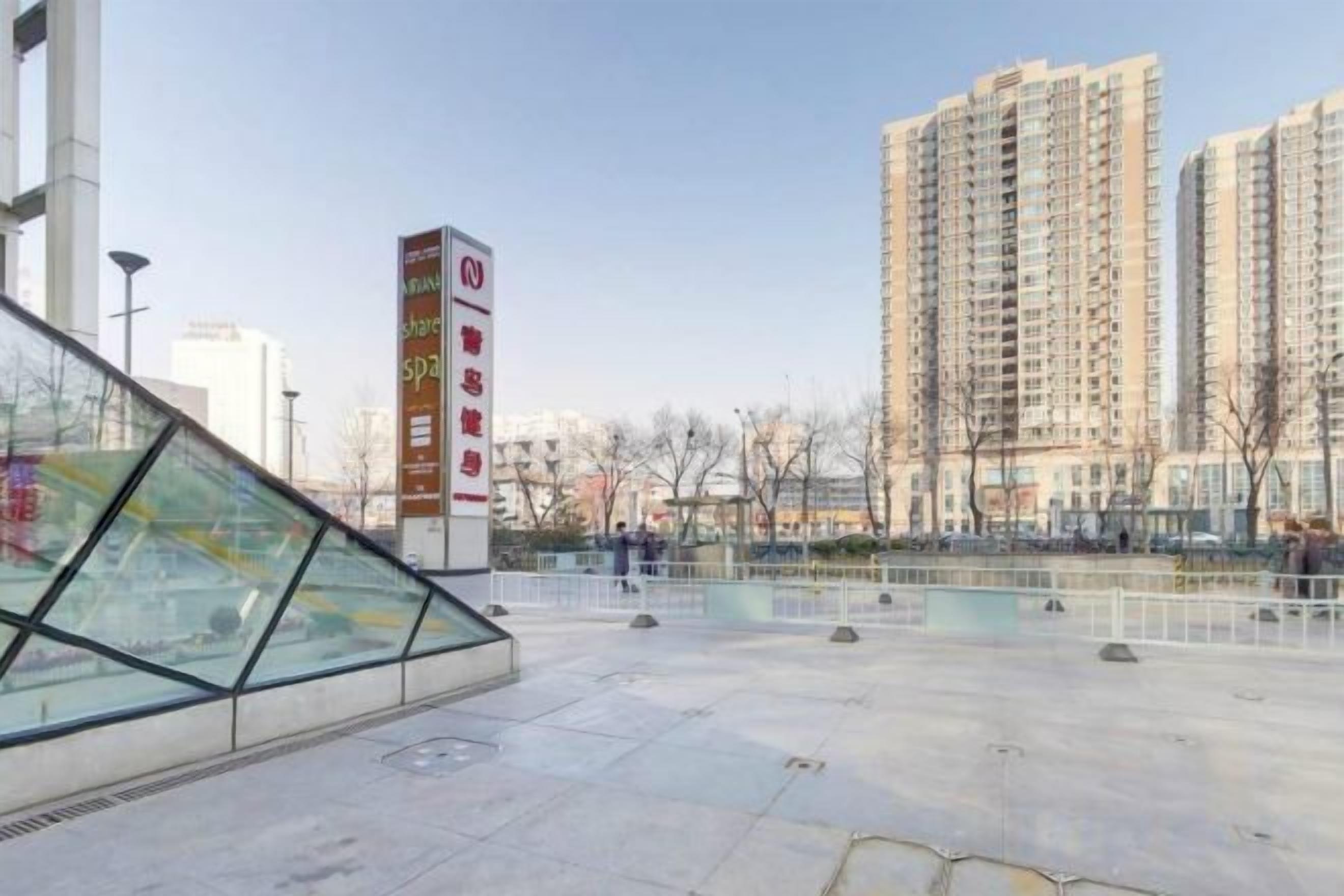 Een modern gebouw met een glazen gevel, een openbaar plein met een fontein, en hoge woontorens op de achtergrond.