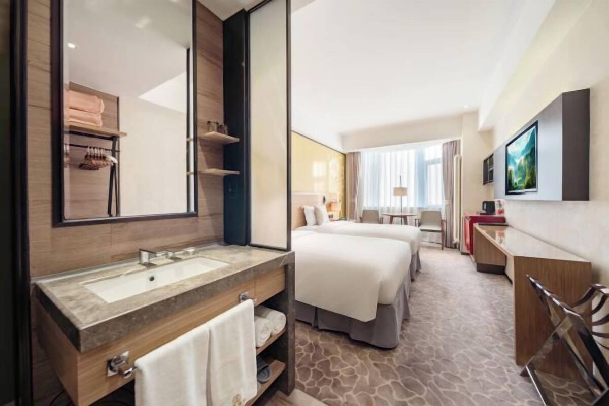 Habitación de hotel con un espejo grande, un lavamanos, dos camas, un escritorio y un televisor.