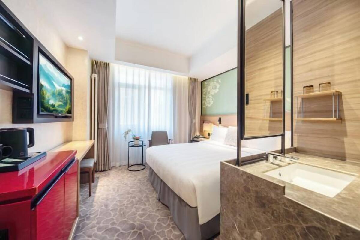 Habitación de hotel con cama, televisión, escritorio con cafetera, pequeña mesa con silla, ventana con cortinas y baño con lavabo y espejo.