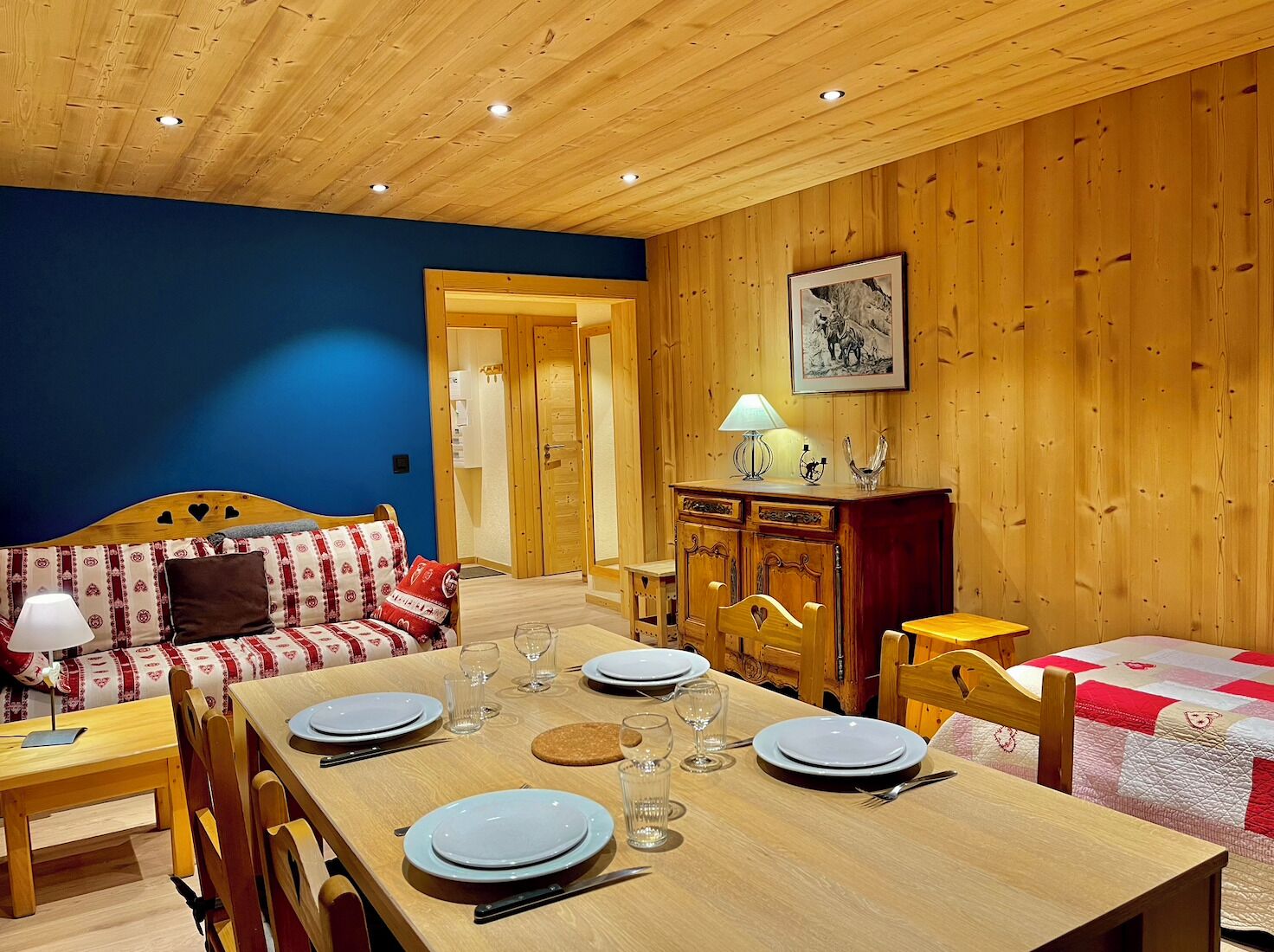 Una habitación acogedora con una mesa de madera dispuesta para cuatro personas, un sofá con cojines estampados y un aparador de madera con una lámpara.