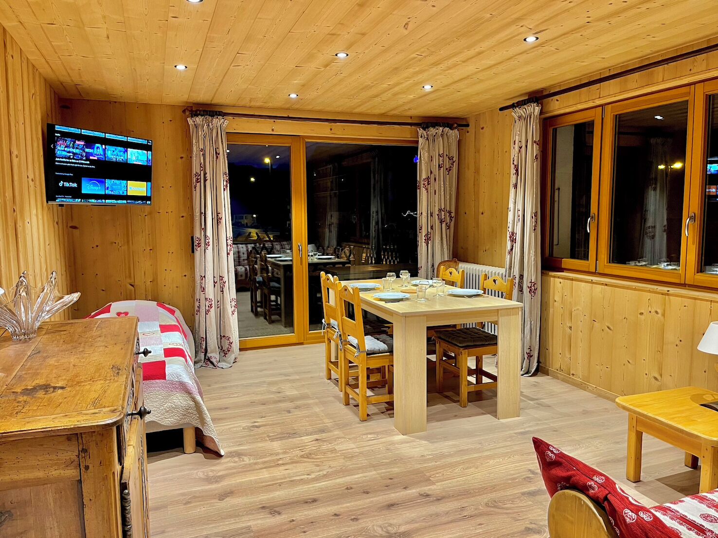 Een houten interieur met een eettafel, stoelen, een aan de muur bevestigde televisie en een glazen schuindeur naar een balkon.