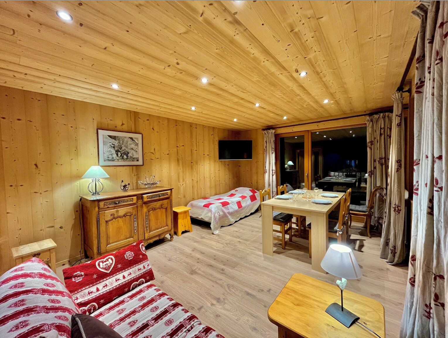 Una habitación acogedora con paredes y techo de madera, una cama con una colcha roja y blanca, un aparador de madera, una mesa con sillas y un televisor montado en la pared.
