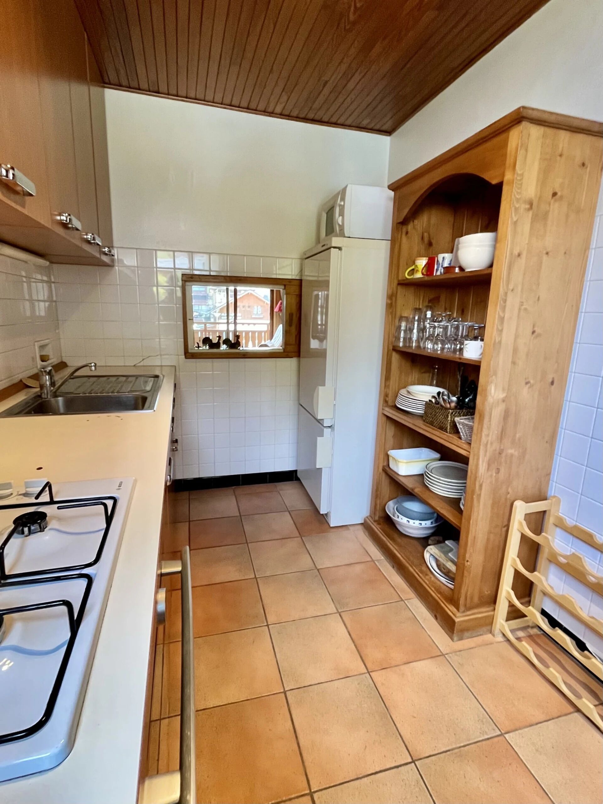 A kitchen with a stove, sink, and wooden cabinets. There is a window with a view of houses outside.