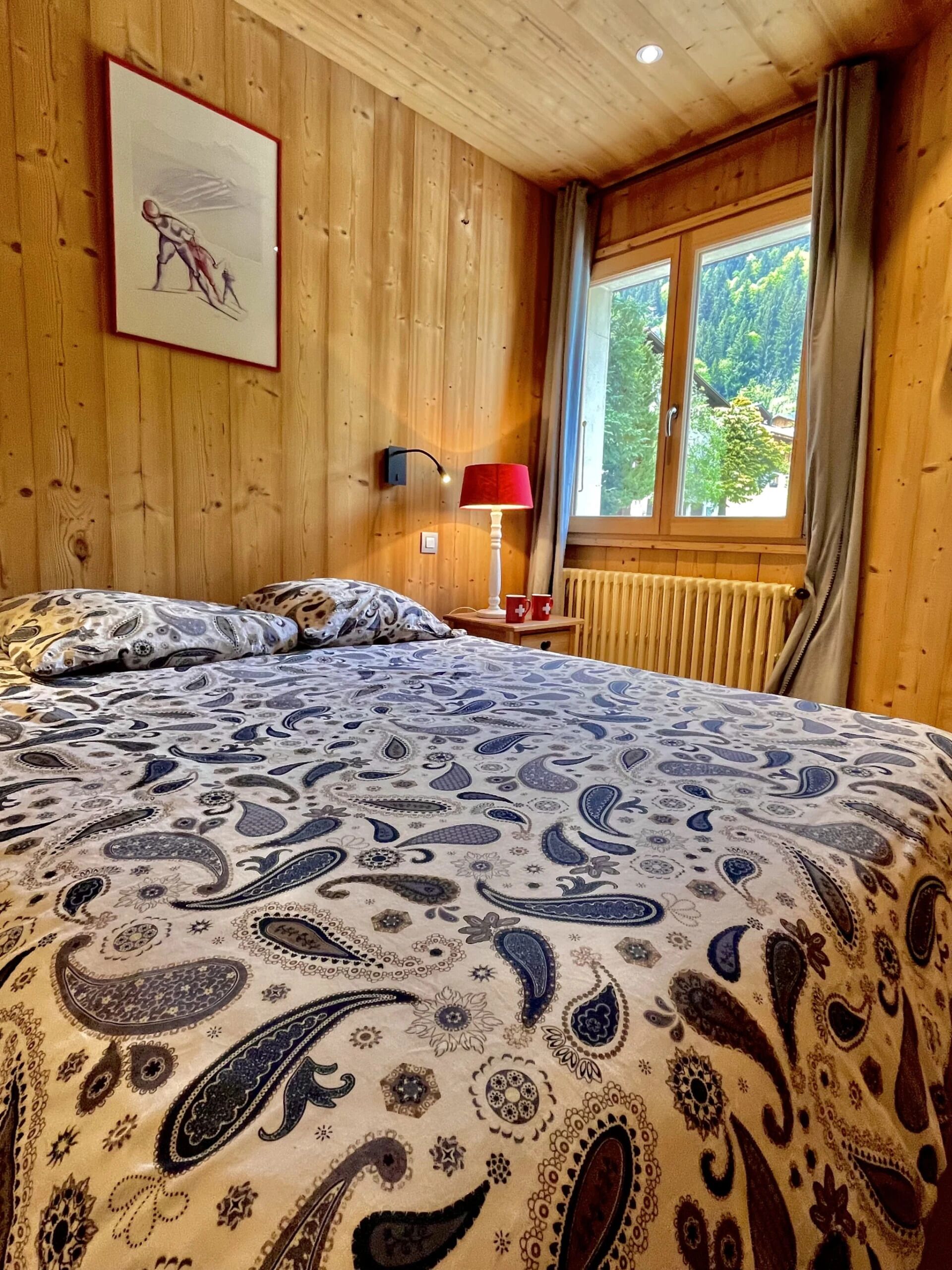 Un dormitorio con paneles de madera, una cama, una mesita de noche, una lámpara y una ventana con vista a la vegetación.