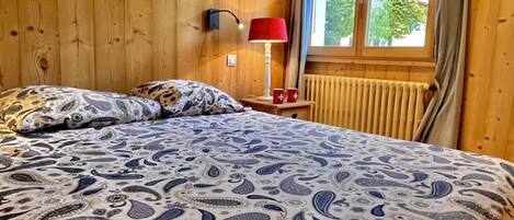 Un dormitorio con paneles de madera, una cama, una mesita de noche, una lámpara y una ventana con vista a la vegetación.