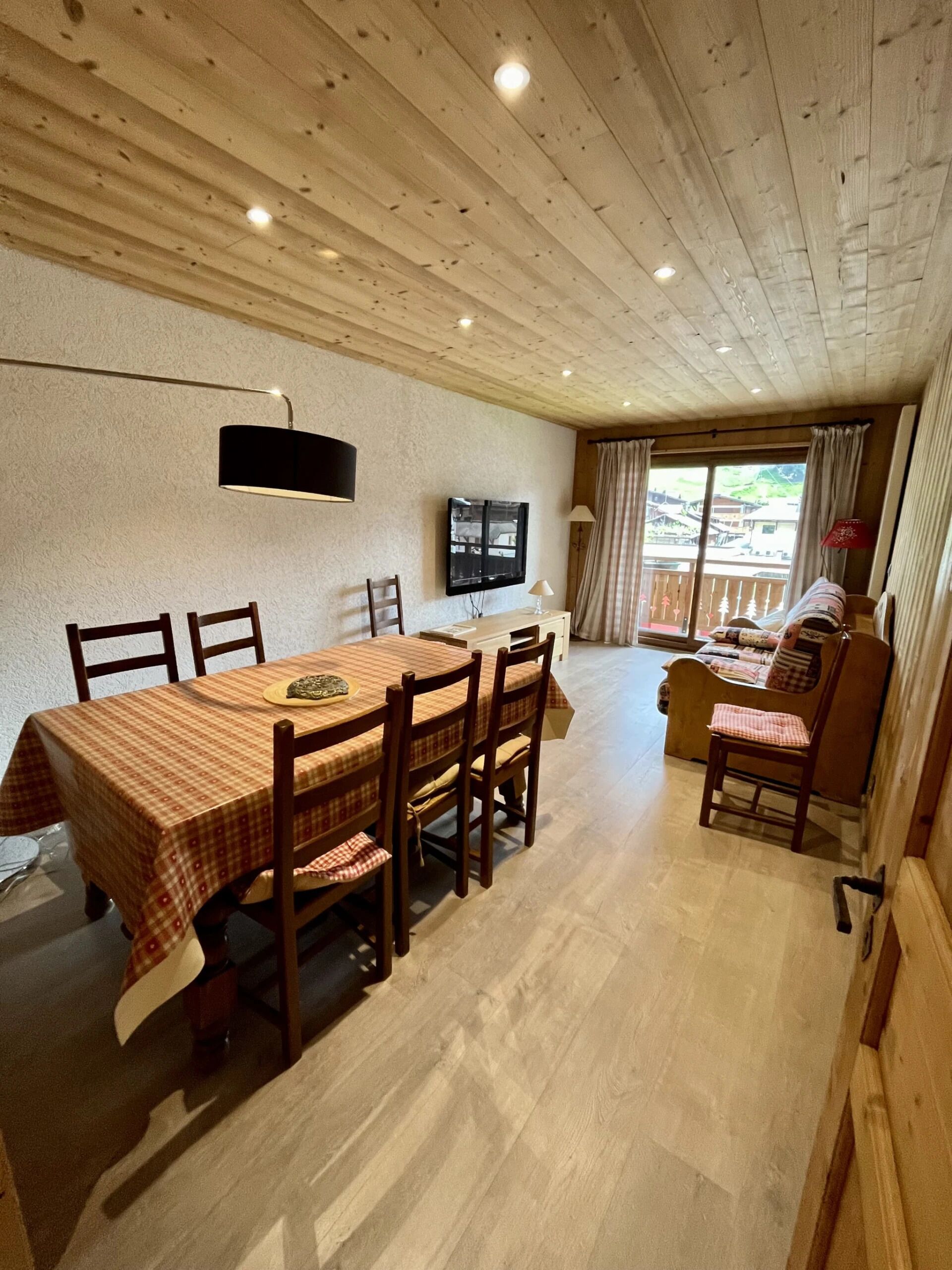 A dining area with a wooden table and chairs, a television mounted on the wall, and a view of the outside through large windows.