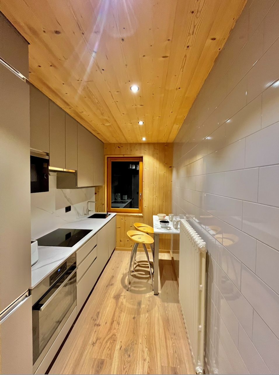 A narrow kitchen with wooden ceiling, white cabinets, a built-in oven, a sink, and stools.