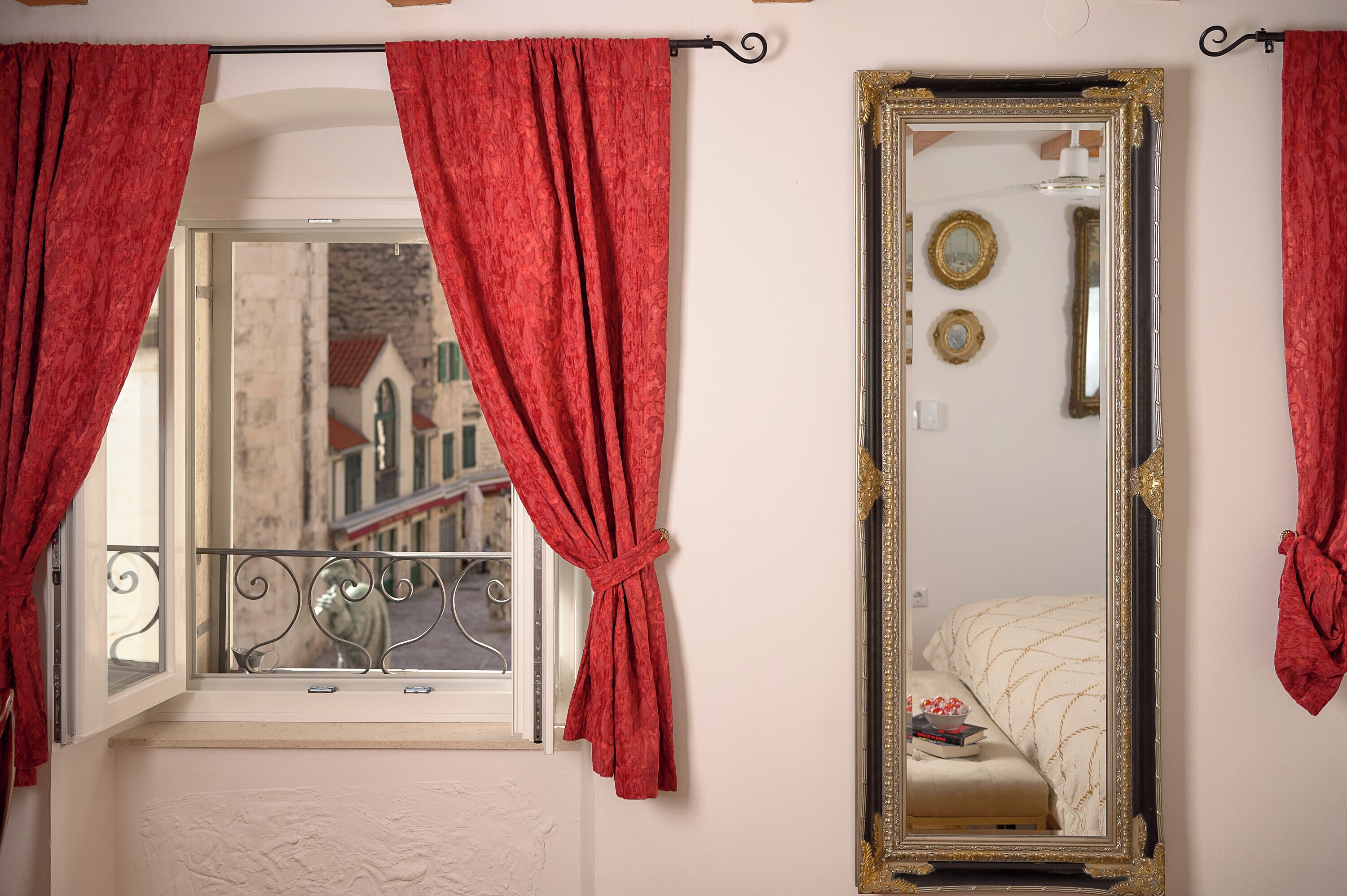 A room with a window, red curtains, a mirror with a gold frame, and a balcony railing.