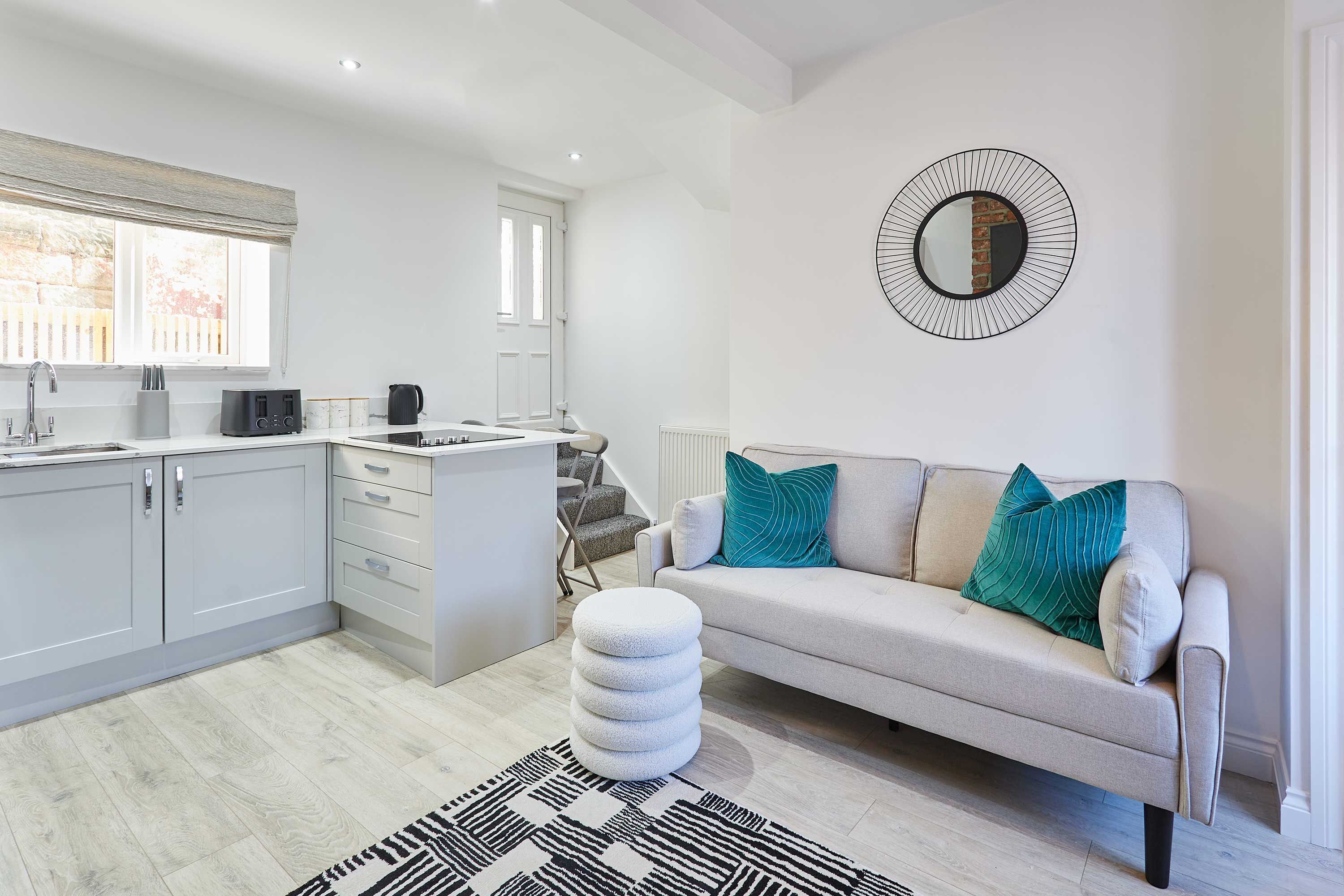 A modern kitchen with white cabinets, a built-in oven, and a dining area with a white table and chairs. There is a white sofa with teal pillows and a black and white rug on the floor.