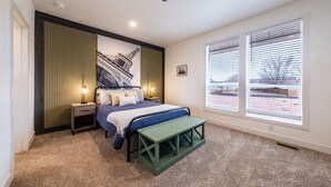 Un dormitorio con una cama, mesitas de noche, un banco, un ventanal grande y un cuadro colgado en la pared.