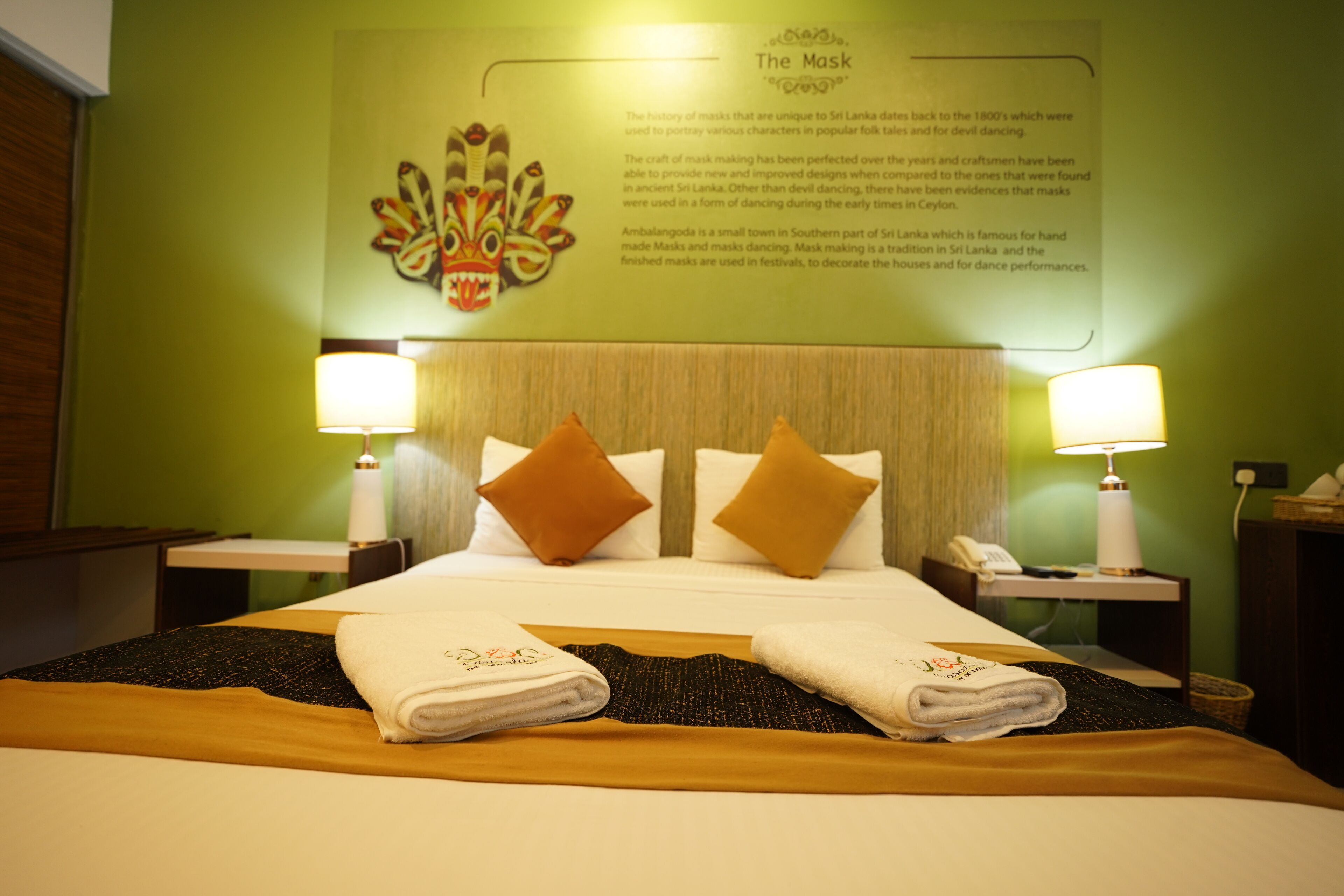 Quarto de hotel com cama bem arrumada, dois travesseiros, luminária fixada na parede e um cartaz informativo emoldurado.