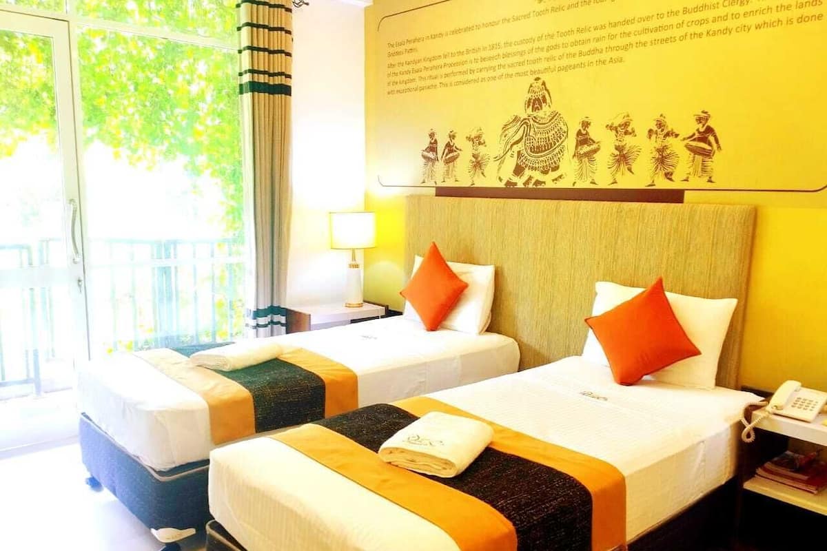 Habitación de hotel con dos camas, cada una con colchón blanco, almohadas naranjas y cabecero marrón. Hay una mesita de noche con lámpara y teléfono, y una ventana con vistas a zonas verdes.