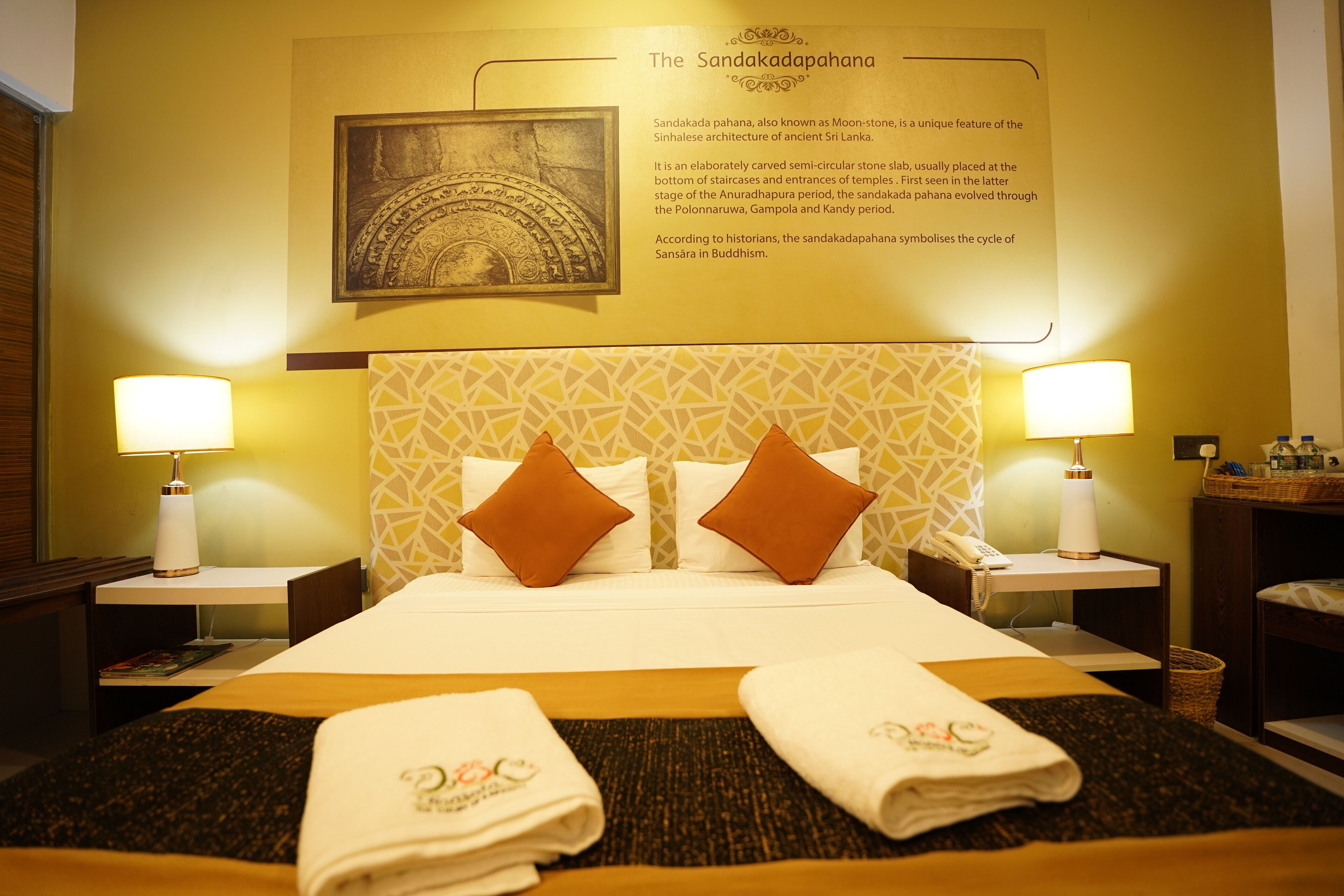 A hotel room with a bed, two nightstands, a wall-mounted lamp, a framed picture, and a description of the Sandakadapahana.