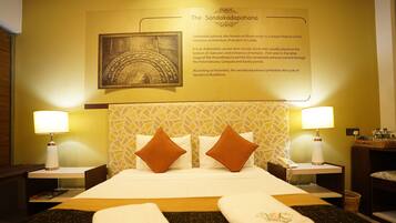 Quarto de hotel com uma cama, duas mesinhas de cabeceira, uma luminária fixada na parede, um quadro e a descrição de Sandakadapahana.