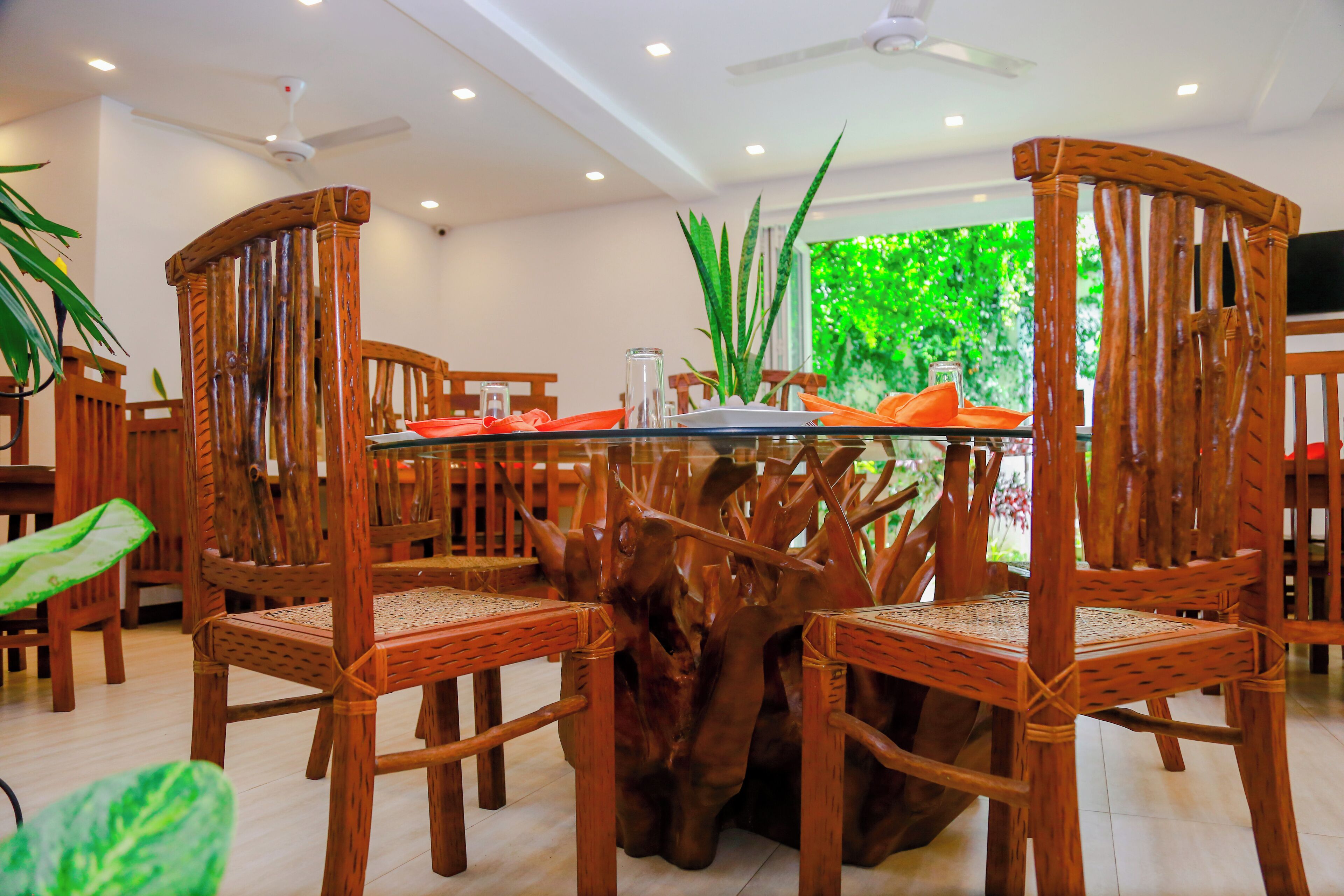 A dining area with wooden chairs and a glass table, surrounded by plants and a view of greenery.