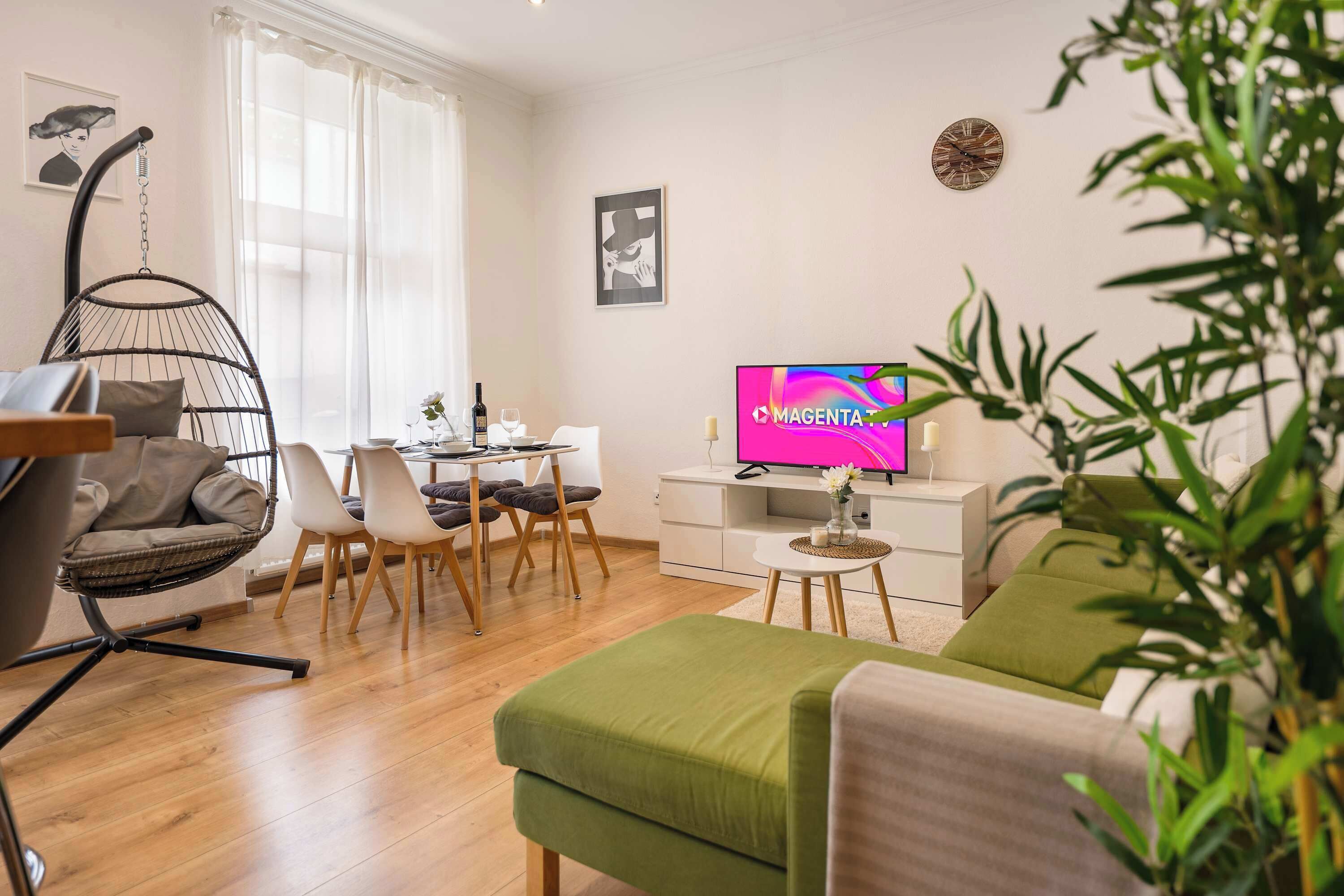 A modern living room with a hanging chair, a dining area, a flat-screen TV, and a green sofa.