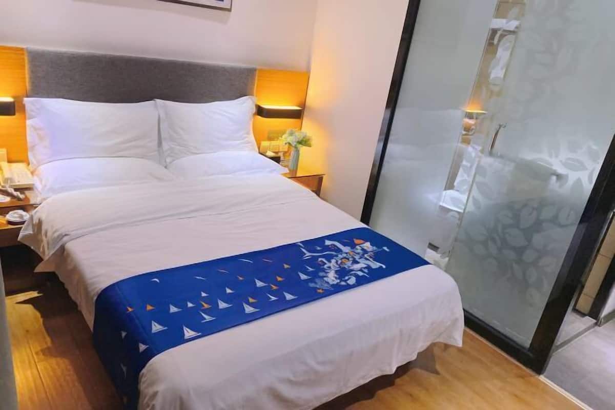 A hotel room with a bed, bedside tables, a nightstand, a decorative blue throw, and a glass partition.