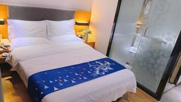 A hotel room with a bed, bedside tables, a nightstand, a decorative blue throw, and a glass partition.