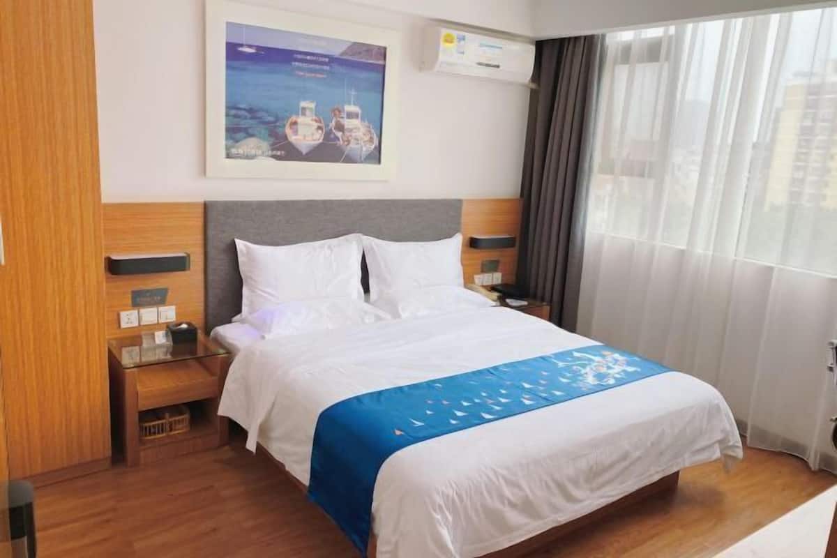 A hotel room with a large bed, a nightstand, a bedside table, a wall-mounted air conditioner, and a window with curtains.