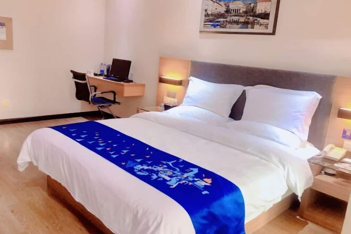 A hotel room with a bed, a desk with a laptop, a chair, a nightstand with a lamp, and a framed picture on the wall.