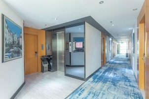Corredor de hotel com piso coberto por carpete, balcão de recepção e porta que dá para outro quarto.