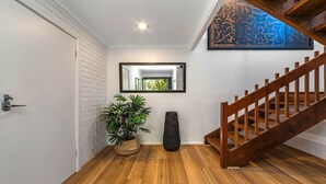 A hallway with a white brick wall, a wooden floor, a potted plant, a black vase, a mirror, and a wooden staircase.