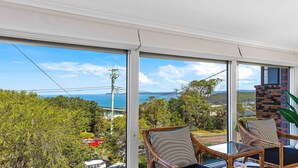 A balcony with wicker chairs and a glass table, overlooking a coastal landscape with trees and a view of the sea.