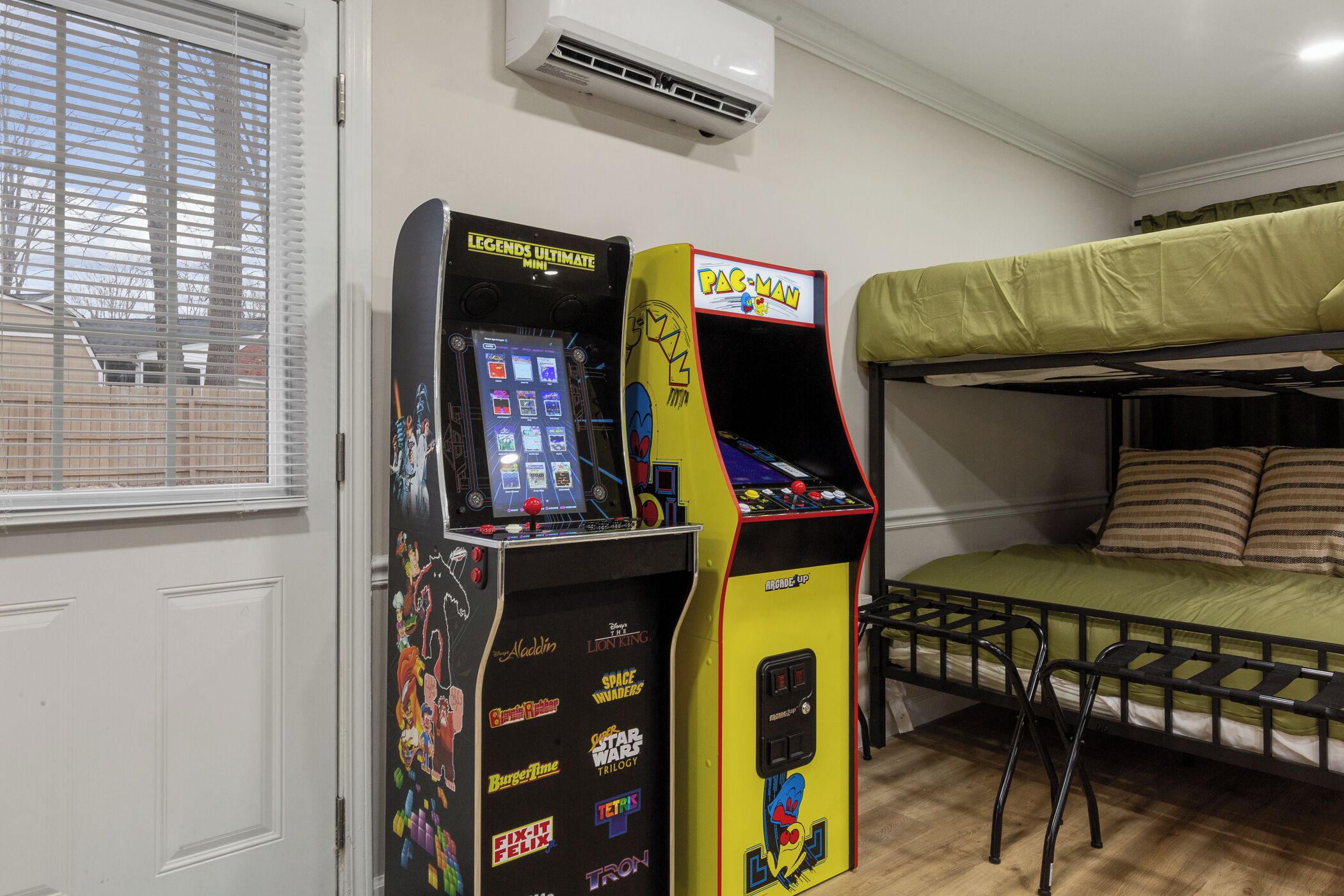 A room with a bunk bed, two arcade machines, and a window with blinds.