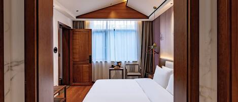 Una habitación de hotel con una cama grande, suelo de madera, un escritorio con silla, una ventana con cortinas e iluminación empotrada en el techo.