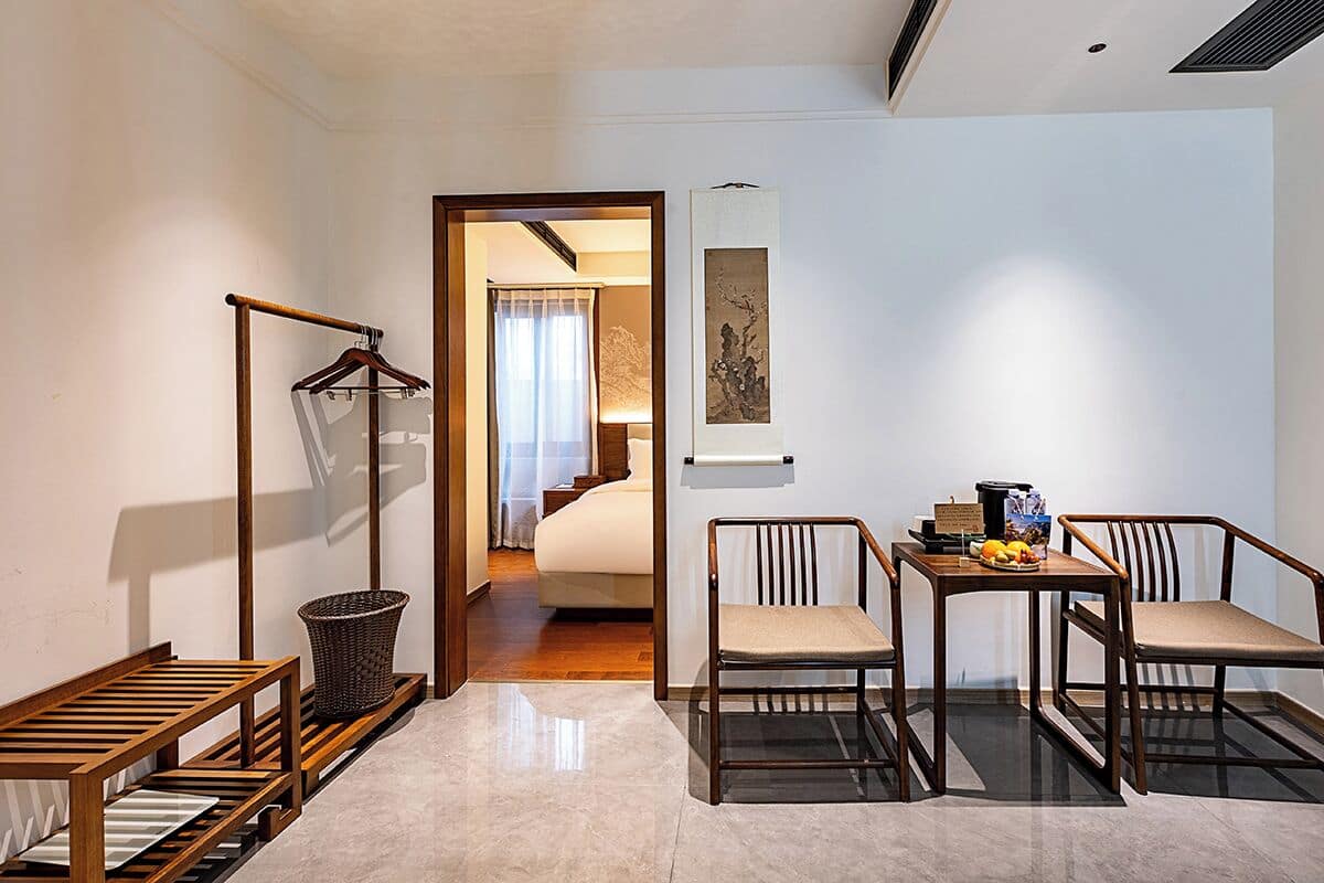 Habitación de hotel con una silla de madera, una mesita con frutas, un perchero y una cama al fondo.