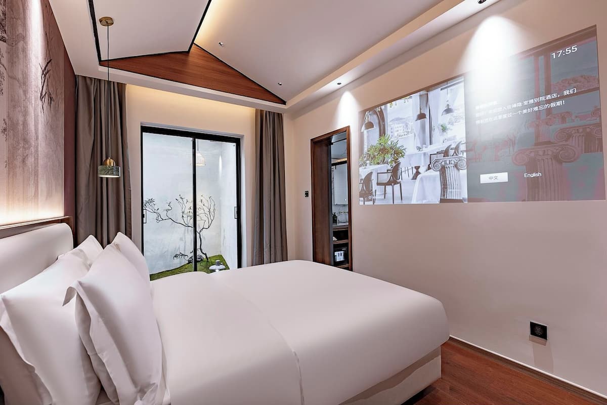 Una habitación de hotel moderna con una cama grande, vistas al exterior, una pantalla de proyección mostrando texto y un diseño de techo de madera.