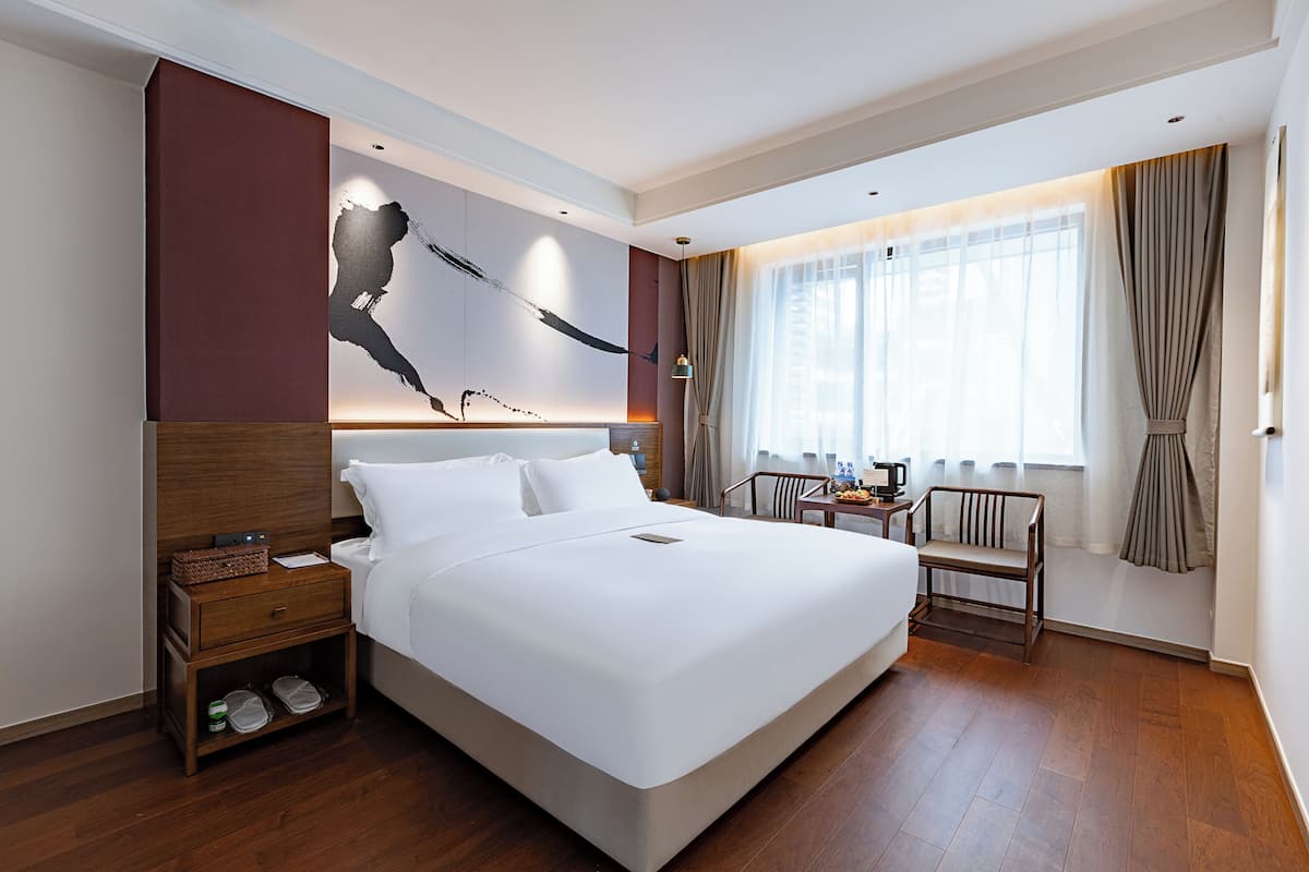 Una habitación de hotel moderna con una cama grande, una mesita de noche de madera, una pequeña mesa con una silla y un ventanal con cortinas.