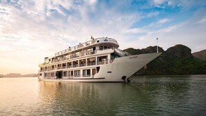 A large, multi-deck river cruise ship with balconies and a flag, docked at a river with a mountainous backdrop. - AE CRUISE HALONG BAY (Tuan Chau)