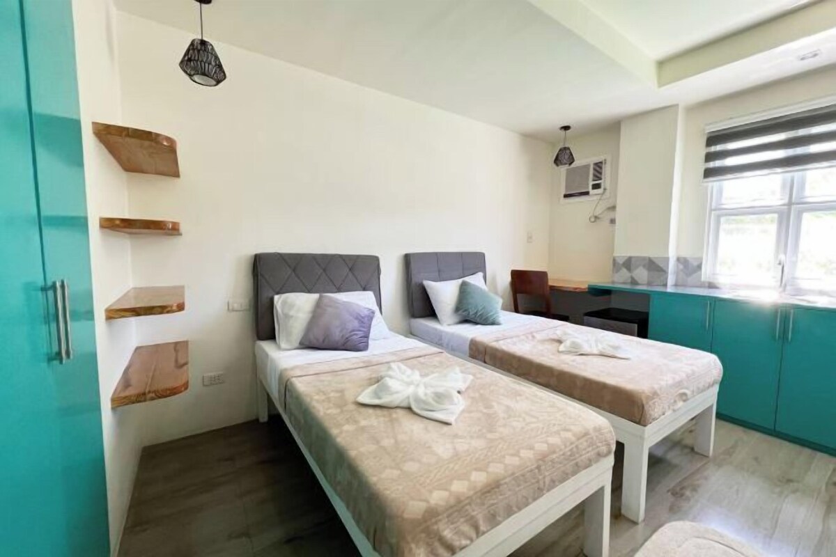 Una habitación con dos camas, una ventana con persianas, un ventilador de techo y una pequeña kitchenette.