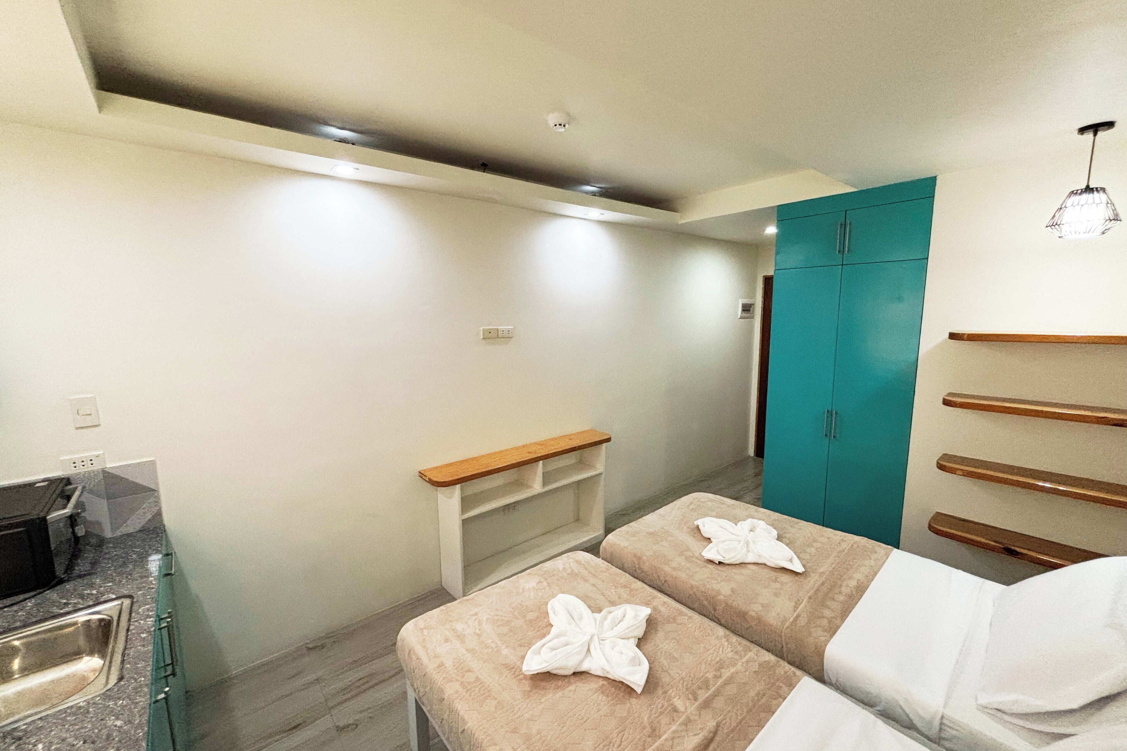 Una habitación de hotel moderna con kitchenette, un armario verde azulado y dos camas con toallas.