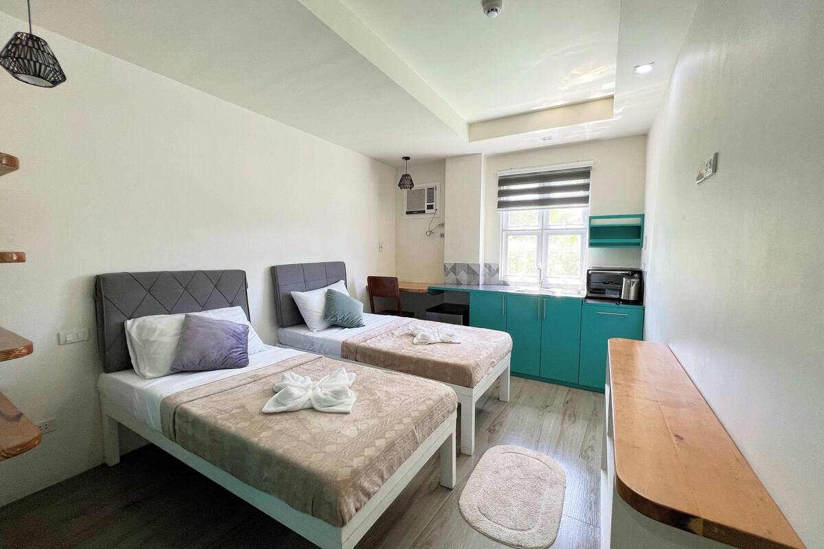 Una habitación pequeña y limpia con dos camas, un banco de madera, una kitchenette y una ventana.