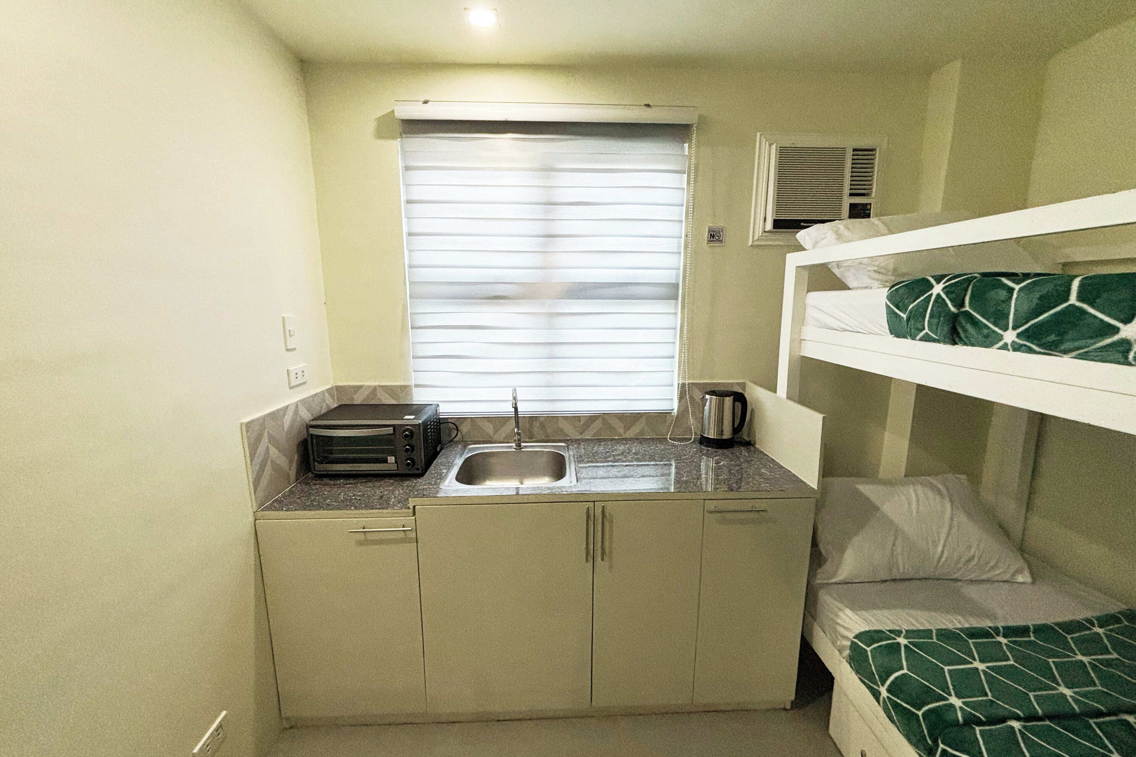 A compact room with a kitchenette, a bed with a green patterned blanket, and a window with blinds.