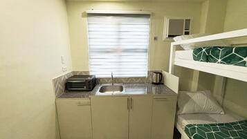 A compact room with a kitchenette, a bed with a green patterned blanket, and a window with blinds.