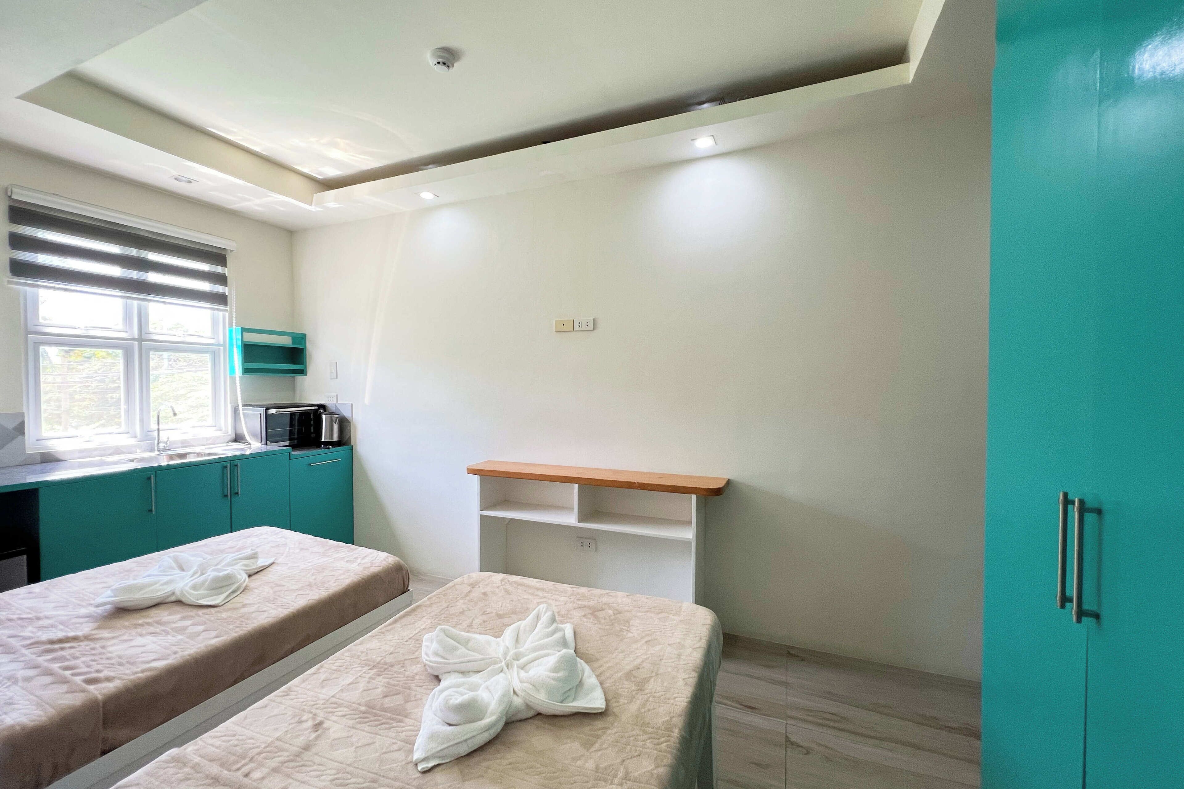 A room with two beds, a window with blinds, a kitchenette, and a teal wardrobe.