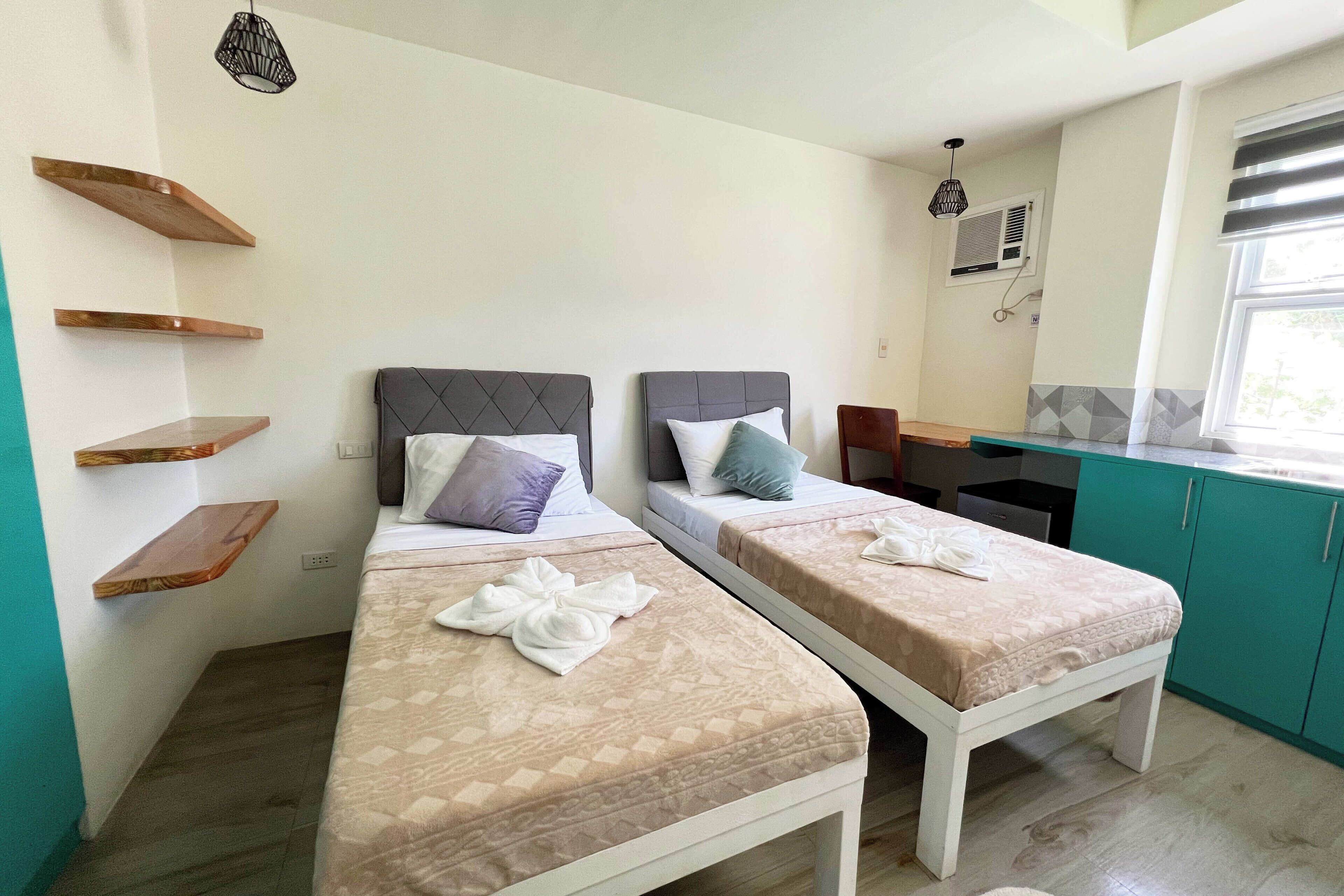 Una habitación con dos camas, estantes de madera, una mesita y un ventanal.