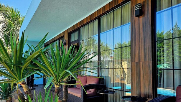 A modern outdoor seating area with wicker furniture, a glass wall, and tropical plants.