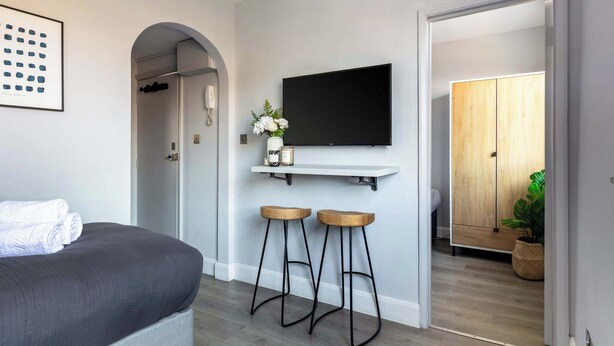 A modern bedroom with a bed, a mounted TV, two stools, and a wooden wardrobe. - Cosy 1BR Steps from Camden Market & Regent's Park (London)