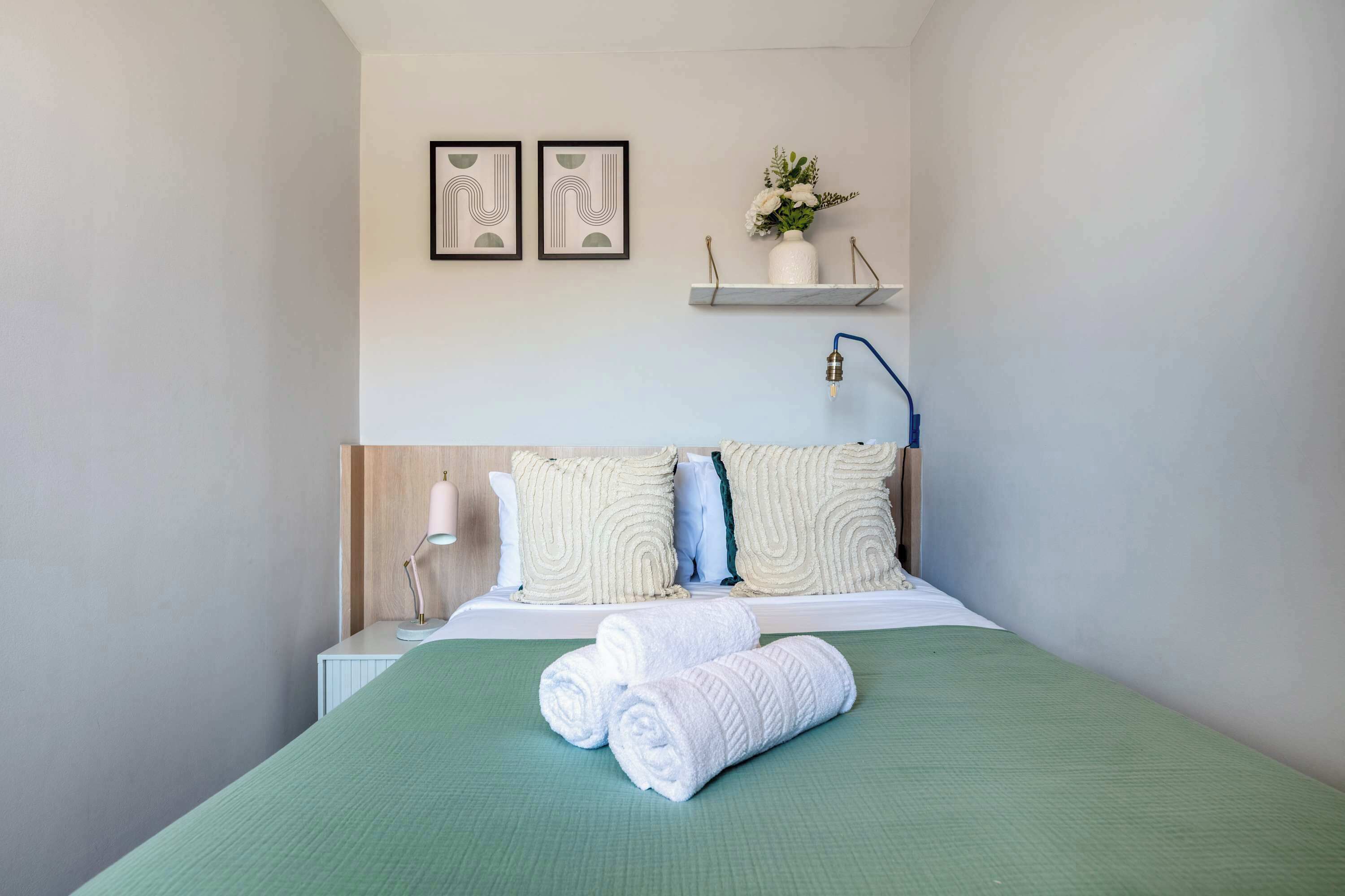 Una cama bien hecha con cubrecama verde, dos almohadas y toallas enrolladas. Hay una lámpara de noche, una mesita de noche pequeña y blanca, y una estantería con una planta.