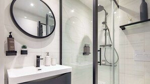 A modern bathroom with a white vanity, a round mirror, and a glass-enclosed shower area. - Cosy 1BR Steps from Camden Market & Regent's Park (London)