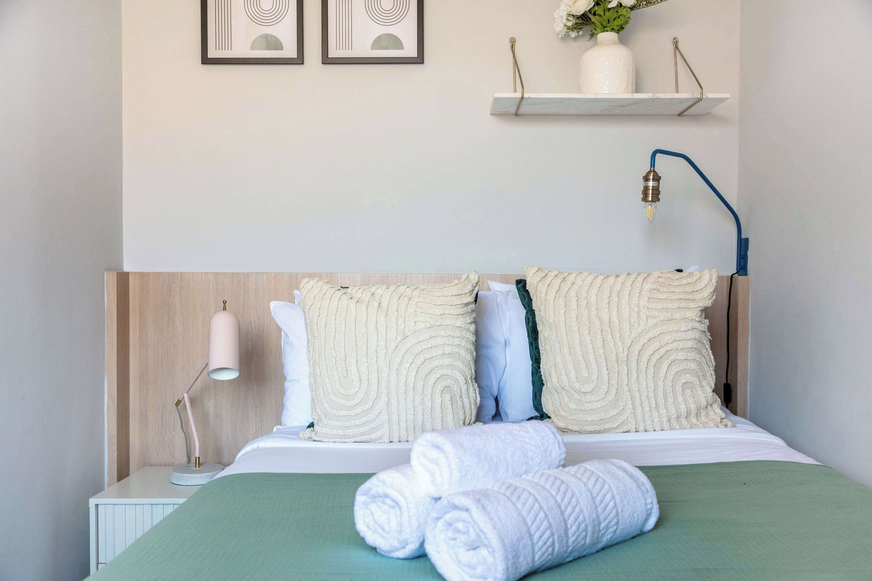 A neatly made bed with rolled white towels, a bedside lamp, and a shelf with a vase and flowers.