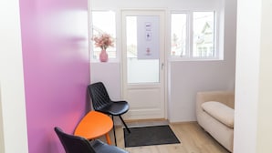 A room with a door, a window, a black chair, an orange chair, a beige sofa, and a vase with dried flowers. - Hostel Fjordur (Hafnarfjörður)