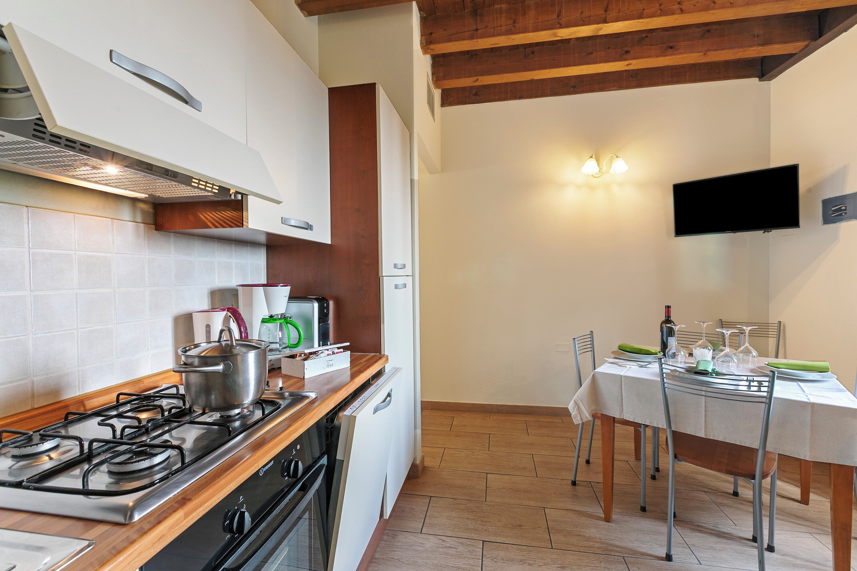 A modern kitchen with a gas stove, oven, and dishwasher. There is a dining table set for two with chairs. A flat-screen TV is mounted on the wall above the table.