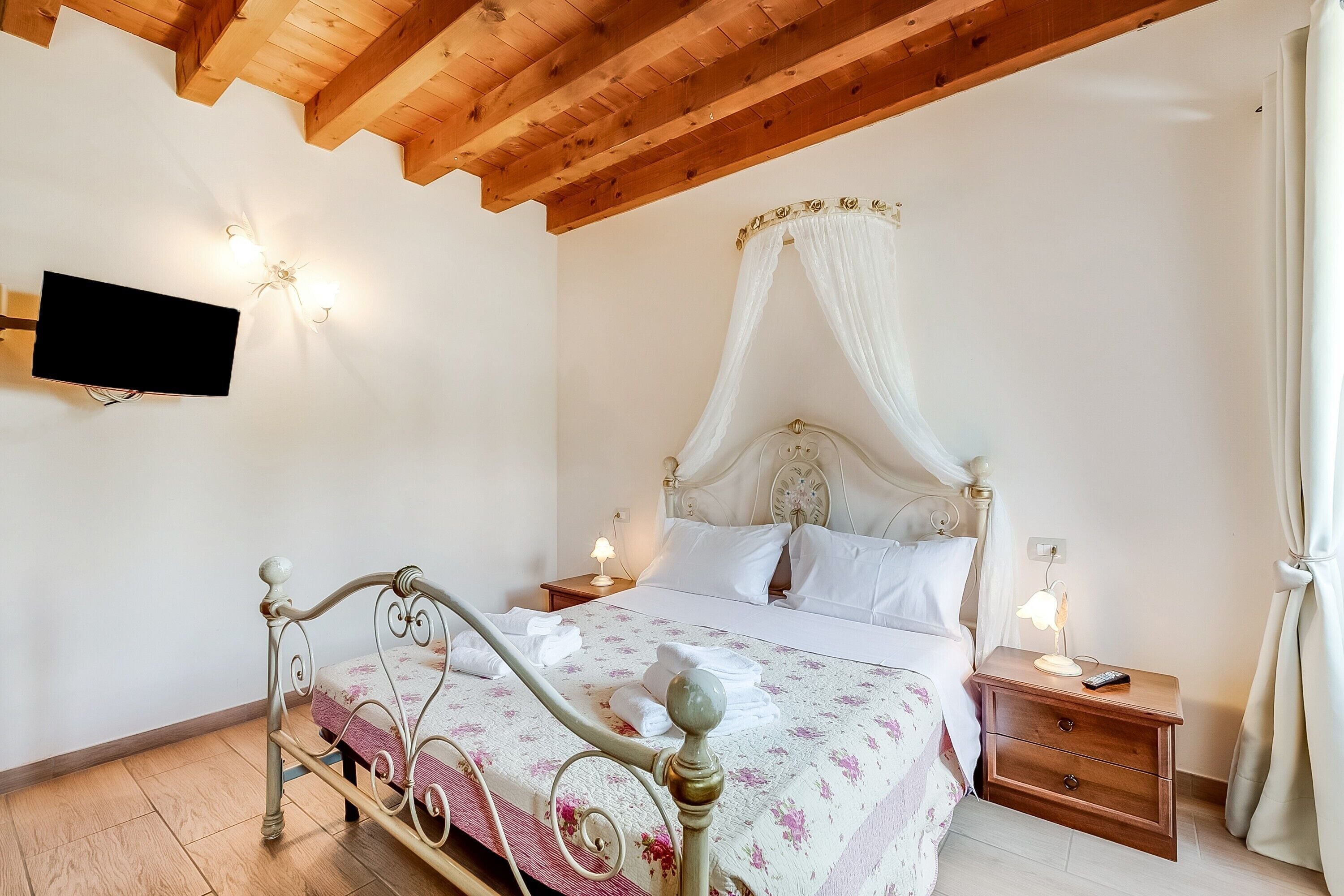 A bedroom with a four-poster bed, a flat-screen TV mounted on the wall, bedside lamps, and a wooden ceiling.